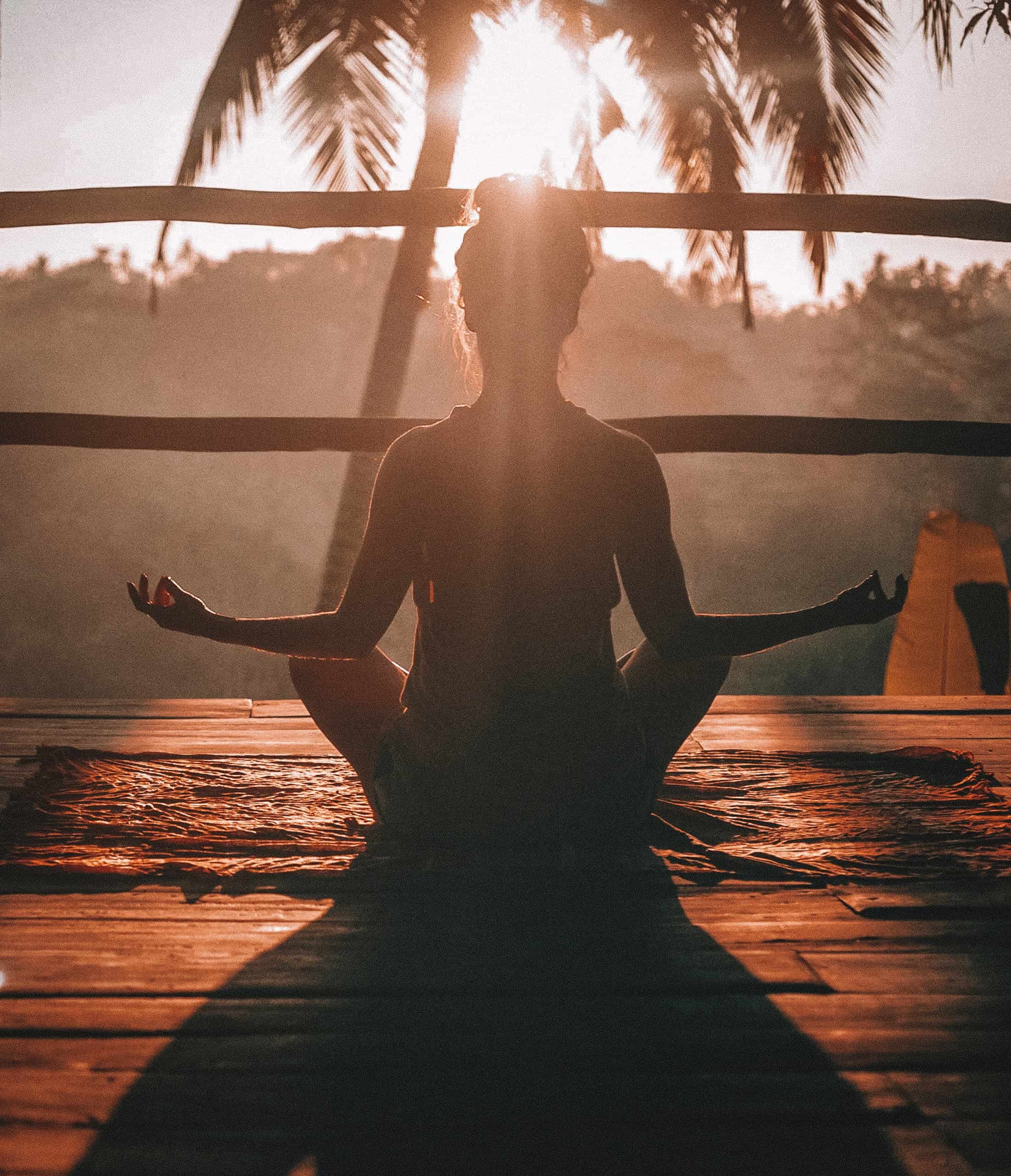 Silhouette of a woman meditating on a wooden platform with her back to the camera and the sunset sunshine in the background